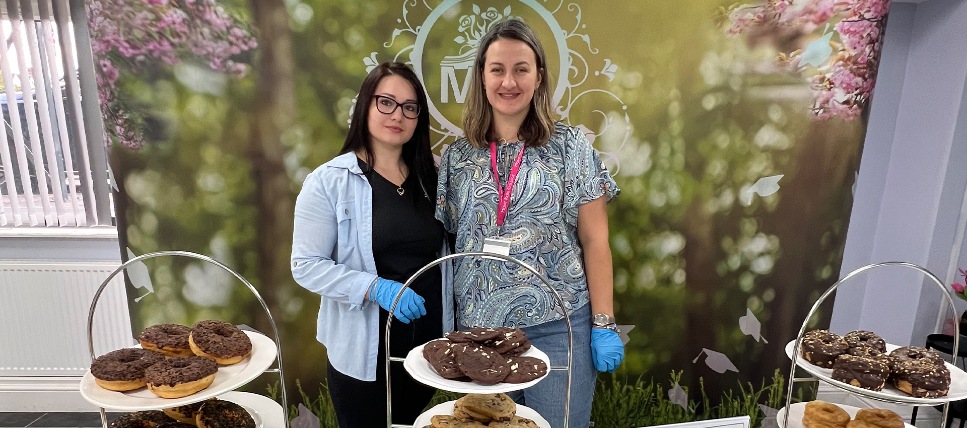 Two staff members posing at the Macmillan Coffee Morning at Mont Rose College, standing behind a dessert display with donuts and cookies, against a floral backdrop.