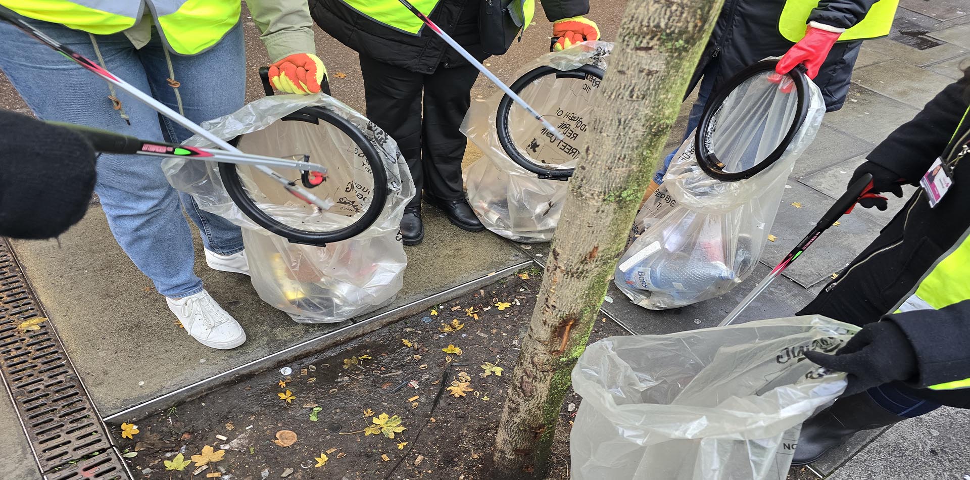 Volunteers wearing high-visibility jackets and gloves use litter pickers to collect trash in transparent bags near a tree during a Greener Ilford Lane litter-picking activity