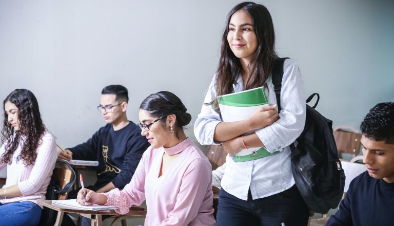 Smiling student standing in a classroom holding books and a backpack Affordability of HND Courses