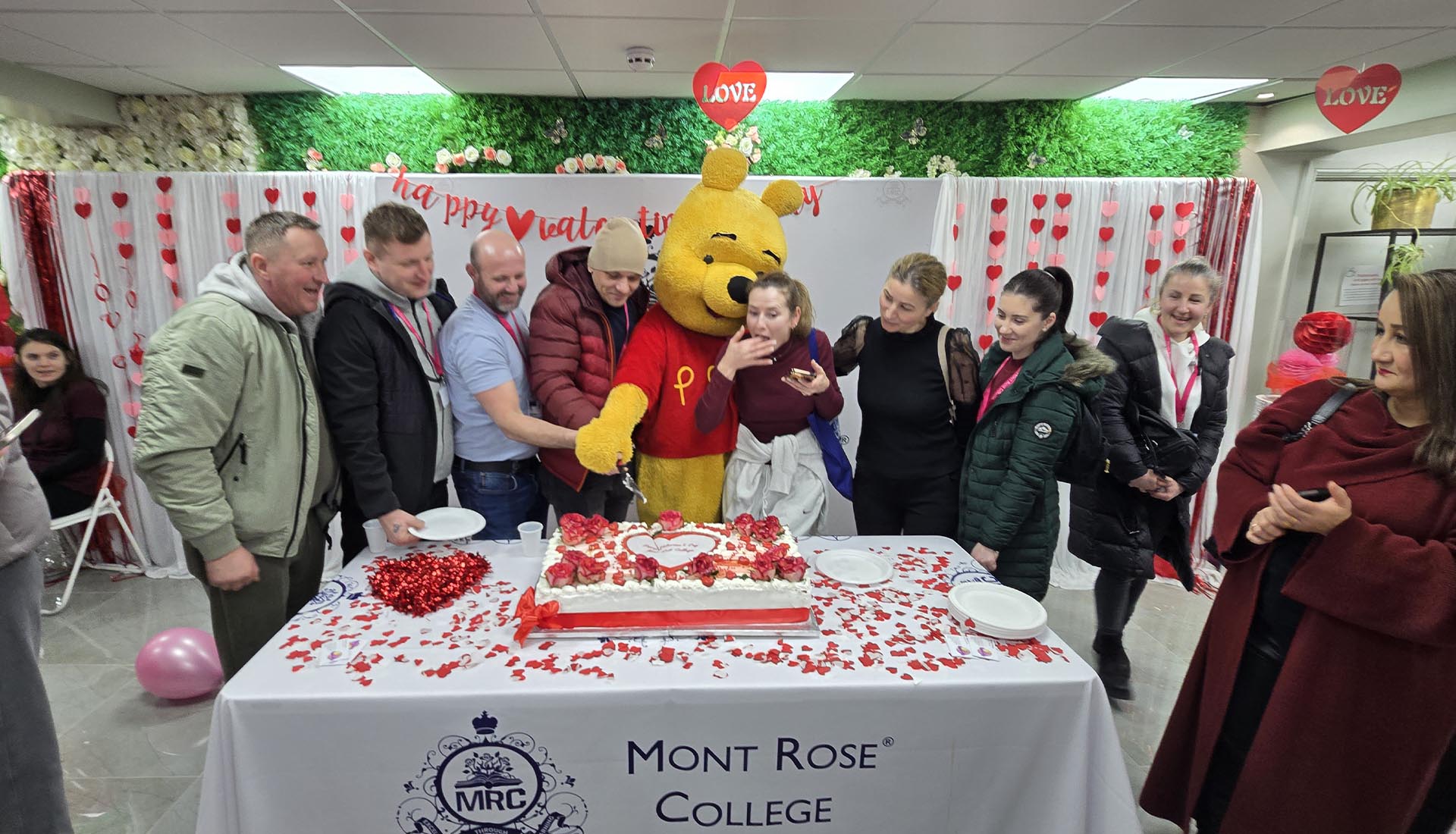 Students and staff celebrating Valentine’s Day at Mont Rose College, cutting a heart-themed cake with a Winnie the Pooh mascot.