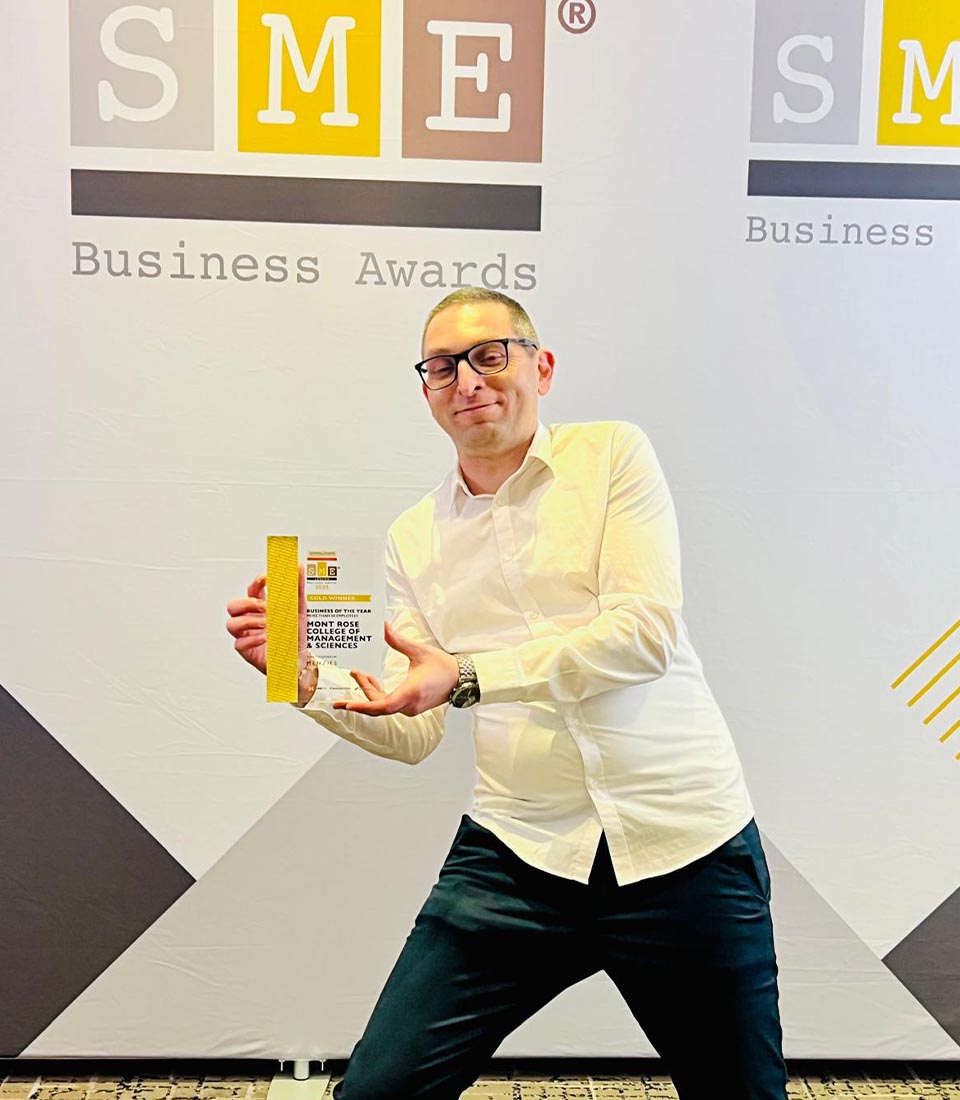 A man in a white shirt proudly poses with an award at the SME London Chamber of Commerce Awards in front of the event backdrop.