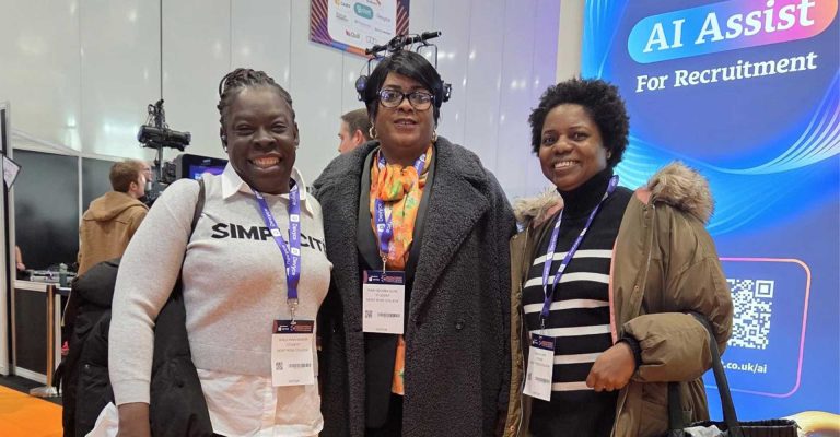 Three women smiling at the Recruitment Agency Expo, wearing visitor badges and standing in front of an AI Assist display.
