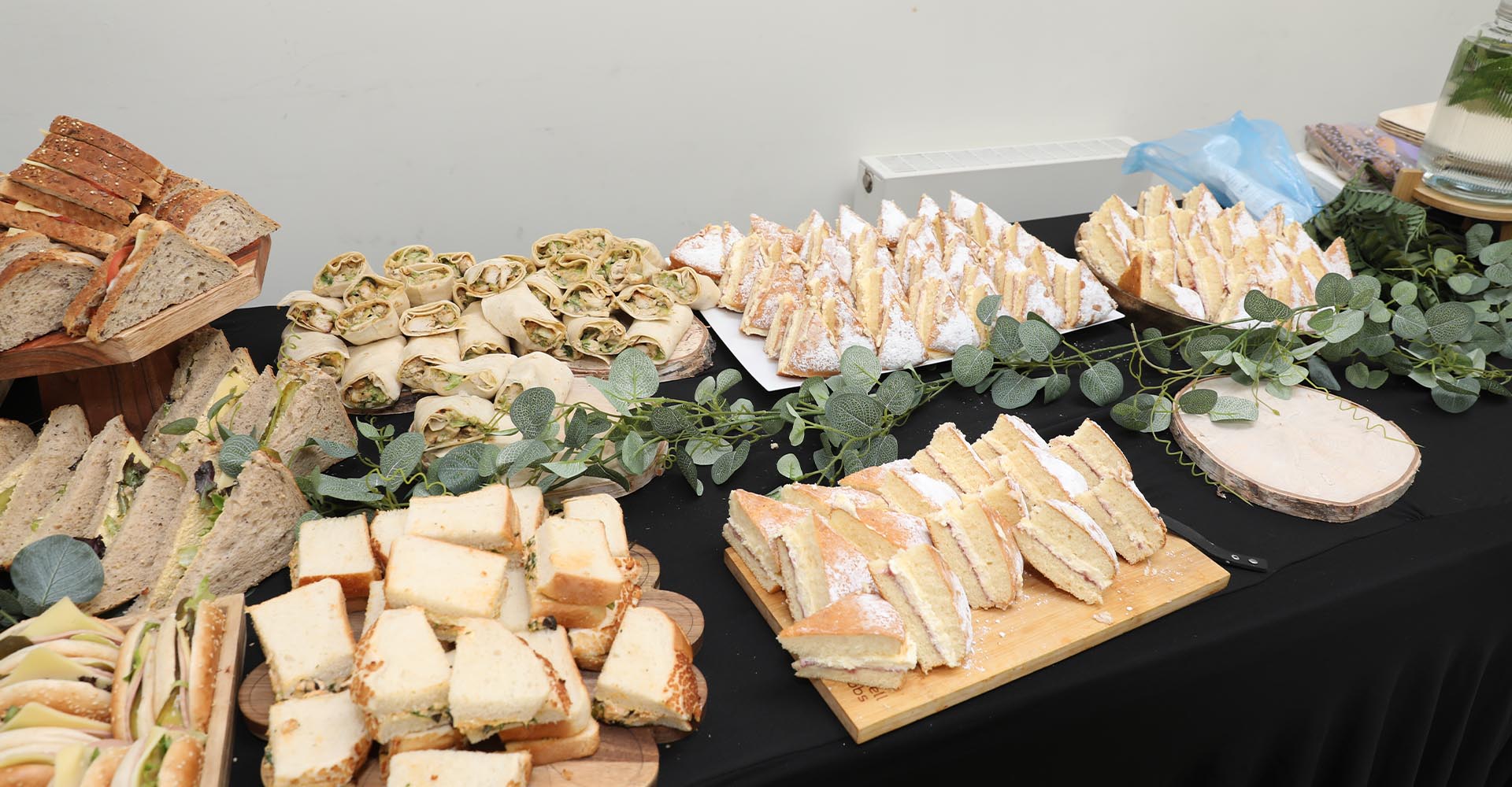 Assorted sandwiches, wraps, and cakes arranged on a black tablecloth for refreshments at the MRC Research Conference 2025.