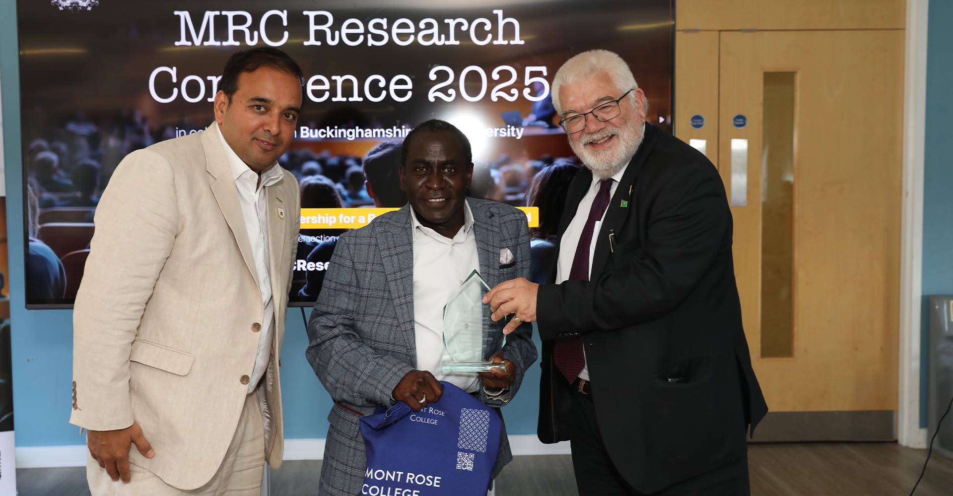 Award recipient posing with two presenters at the MRC Research Conference 2025, holding a trophy and Mont Rose College bag.
