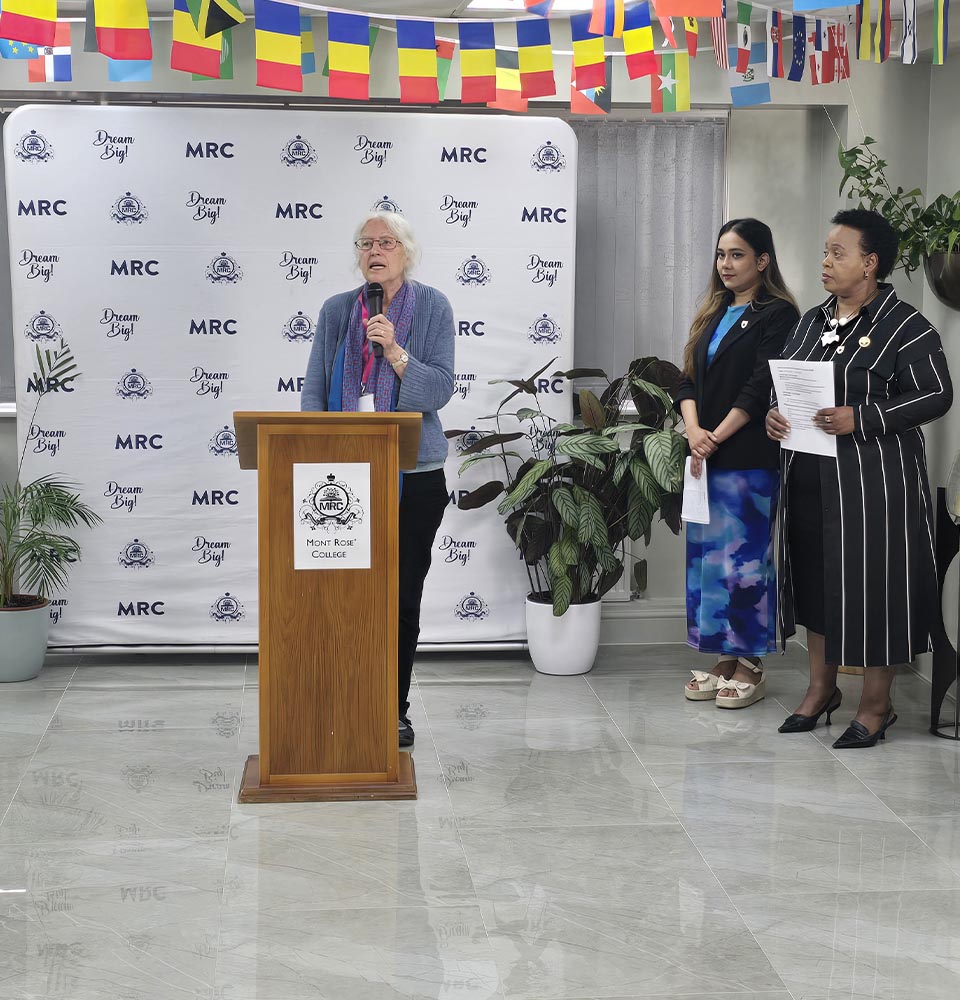 Speaker addressing audience at Mont Rose College podium during Environment Day Programme, with two women standing beside her.