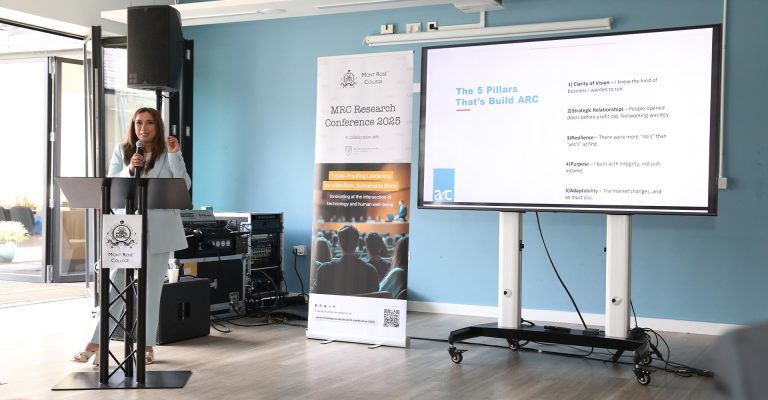 Speaker presenting at Mont Rose College podium during the MRC Research Conference, with a screen showing leadership pillars.