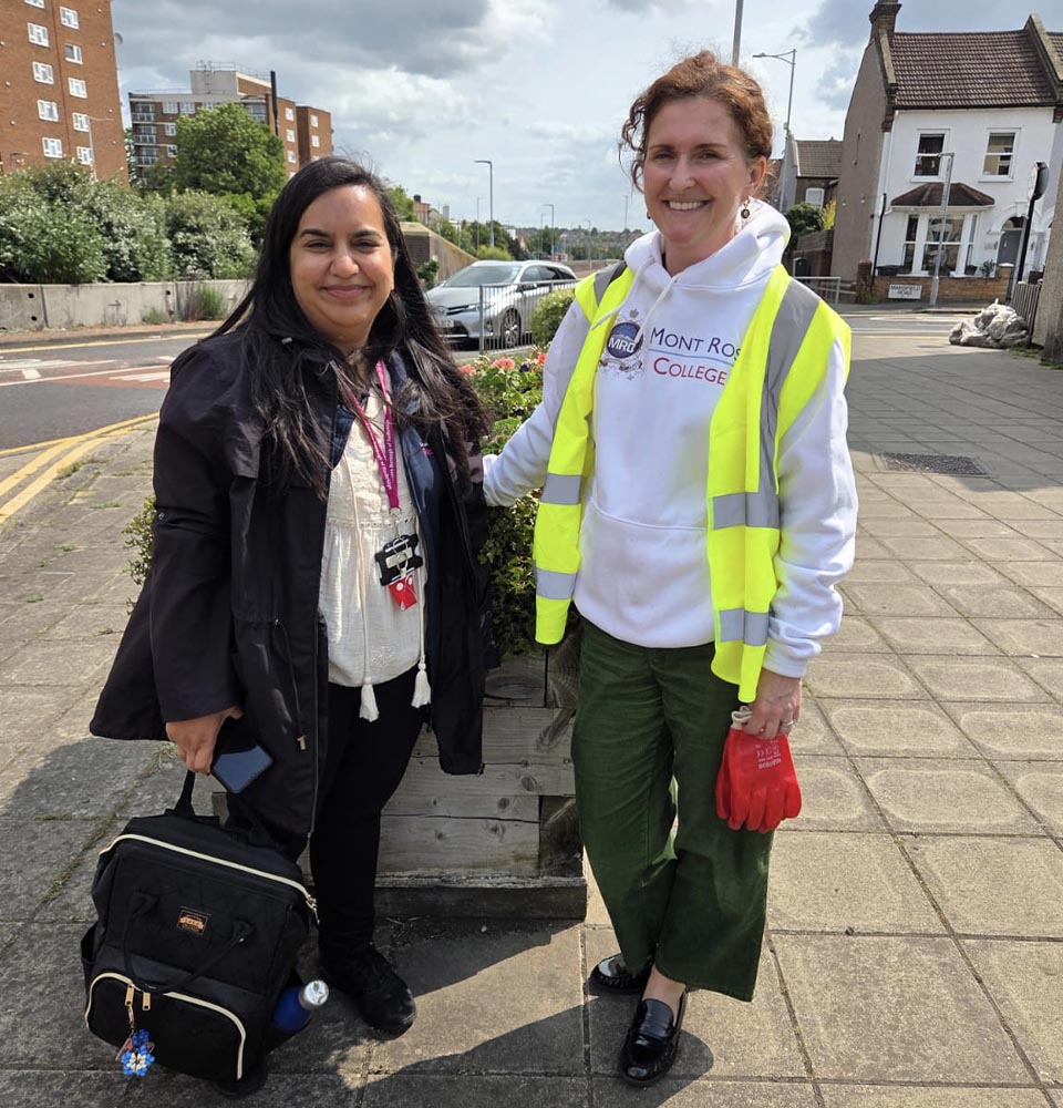 Two women standing on a city sidewalk near plants, participating in the Greener Redbridge community initiative.
