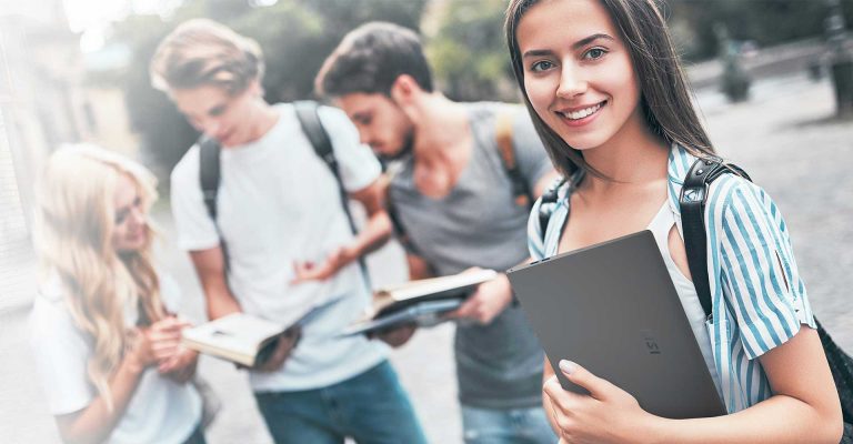 Smiling student holding a laptop, representing HND in Digital Technologies, with classmates studying in the background.
